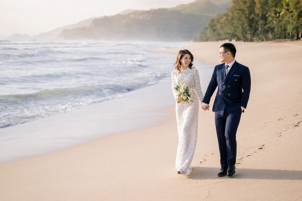 Cô dâu áo dài ren trắng nắm tay chú rể vest xanh than đi dọc mép sóng, phía xa là dãy núi mờ sương