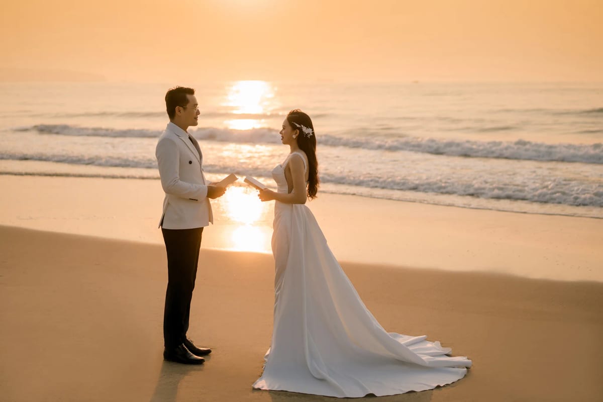 Cô dâu chú rể đứng đối diện đọc lời thề trên bãi cát, vệt nắng bình minh chạy dài giữa hai người