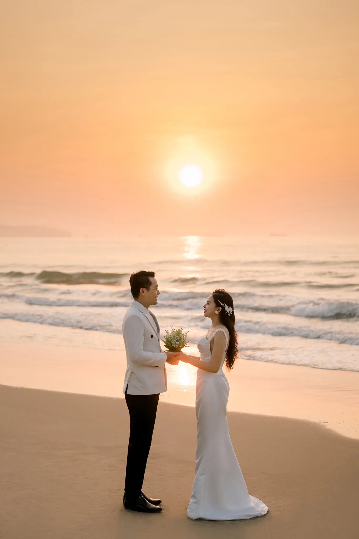 Chú rể vest trắng trao bó hoa cho cô dâu trên bãi biển, mặt trời mọc phía sau in vệt sáng xuống mặt nước