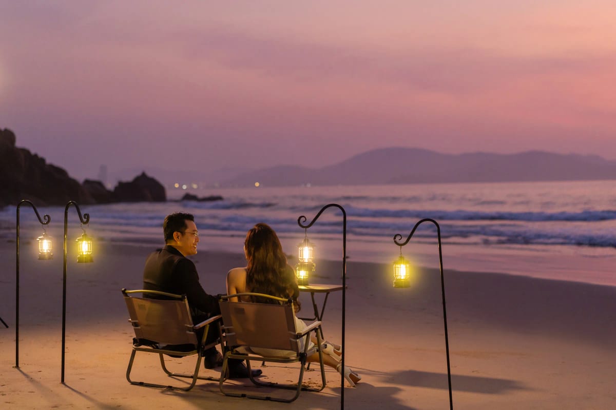 Cặp đôi ngồi trên ghế xếp nhìn ra biển lúc hoàng hôn hồng tím, xung quanh là những chiếc đèn lồng treo
