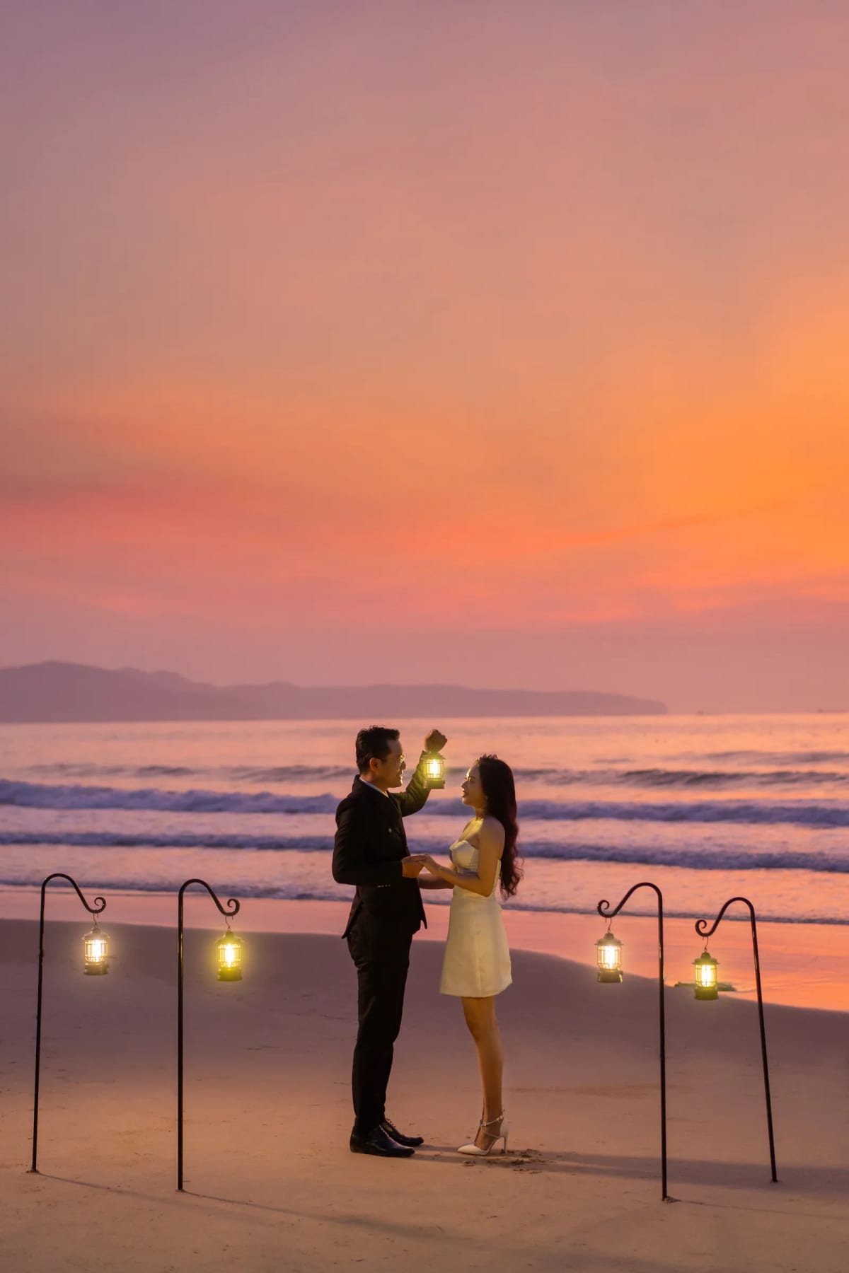 Chú rể nâng chiếc đèn lồng lên ngang tầm mắt cô dâu trên bãi biển lúc trời chuyển tím cam