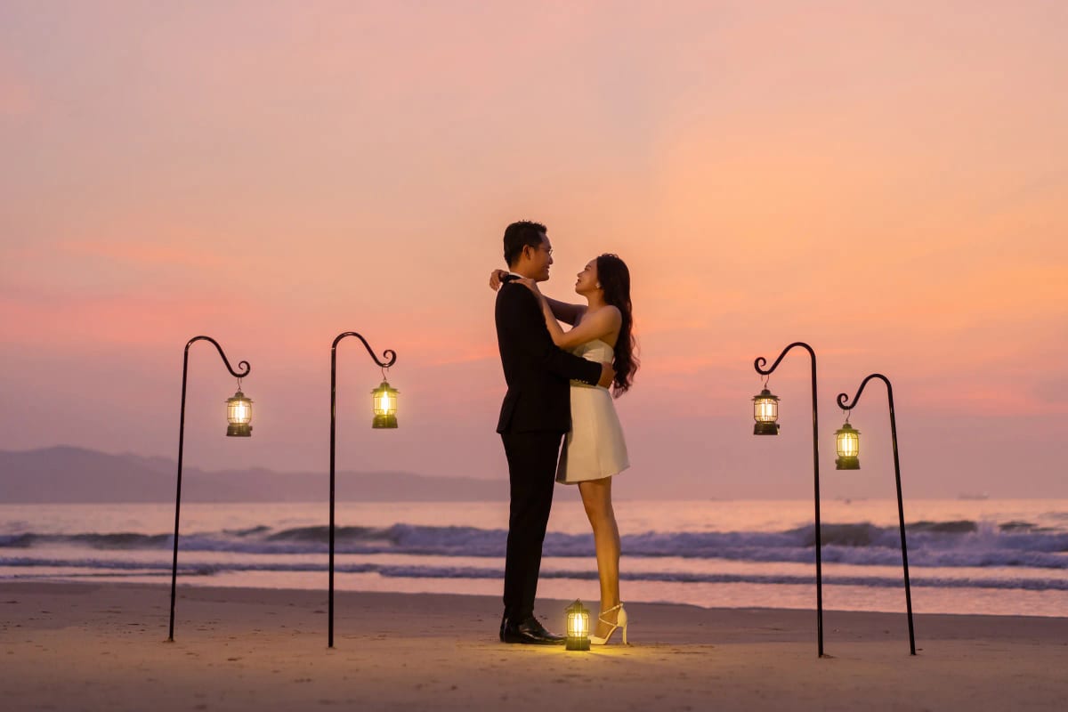 Cô dâu vòng tay ôm cổ chú rể giữa hàng đèn lồng trên bãi cát, nền trời hoàng hôn hồng cam