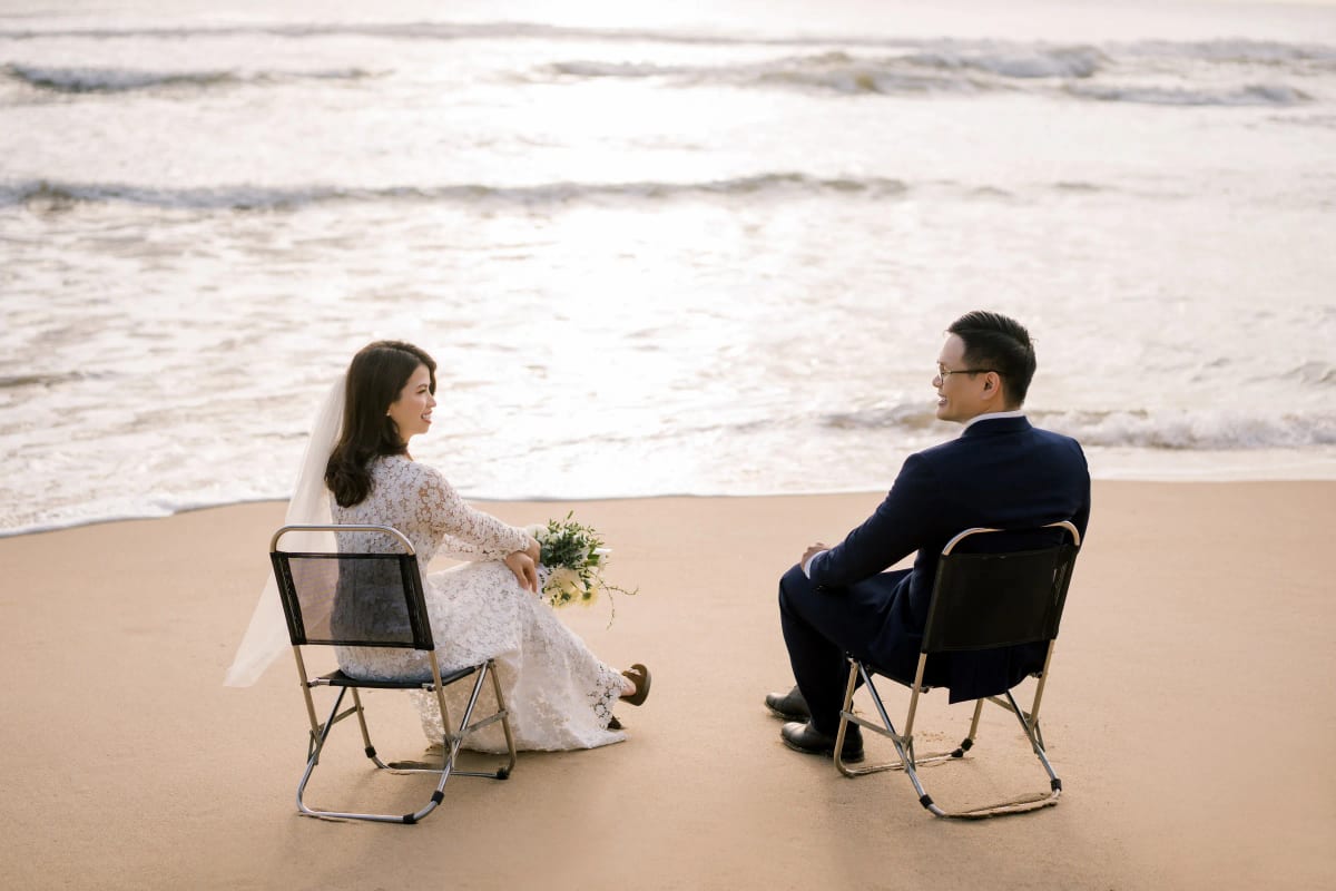 Cô dâu và chú rể ngồi trên hai chiếc ghế xếp quay mặt vào nhau trên bãi biển vắng