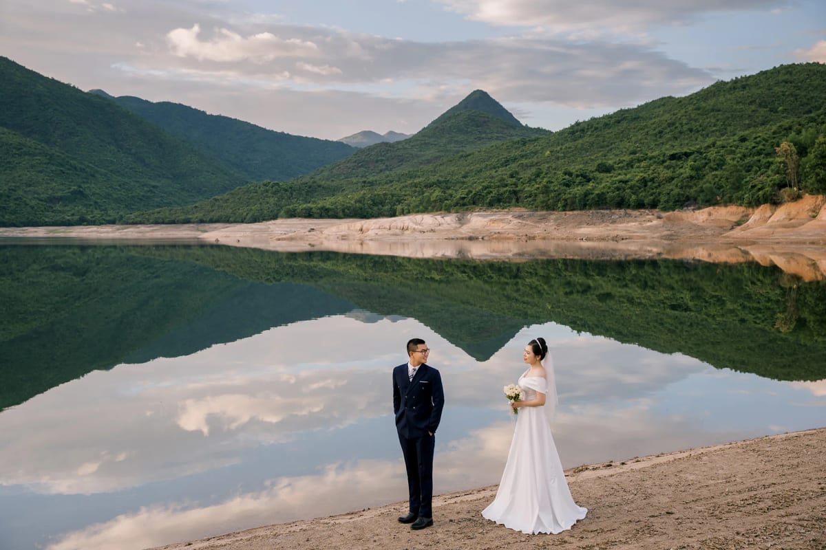 Cô dâu chú rể đứng nhìn nhau bên hồ nước phẳng như gương, dãy núi xanh và mây trắng in bóng phía sau