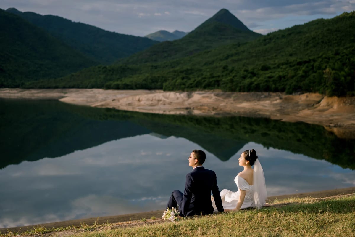 Cô dâu chú rể ngồi tựa lưng vào nhau trên bờ cỏ ven hồ, ngọn núi hình chóp phản chiếu xuống mặt nước