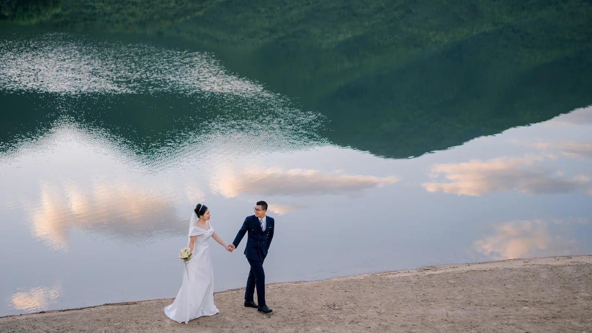 Cặp đôi nắm tay đứng bên hồ nước phẳng lặng phản chiếu mây trời và sườn núi xanh