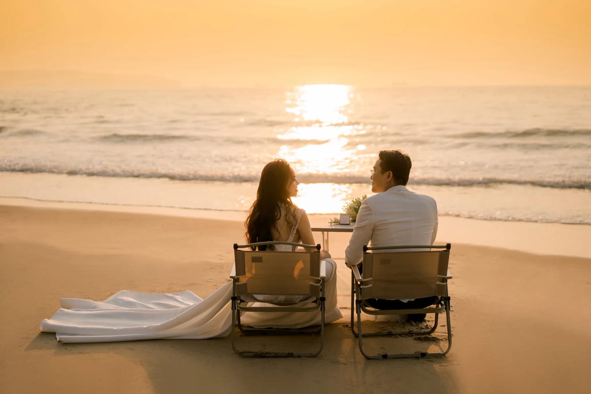 Cô dâu chú rể ngồi bên chiếc bàn nhỏ trên bãi cát nhìn ra vệt nắng vàng trải trên mặt biển