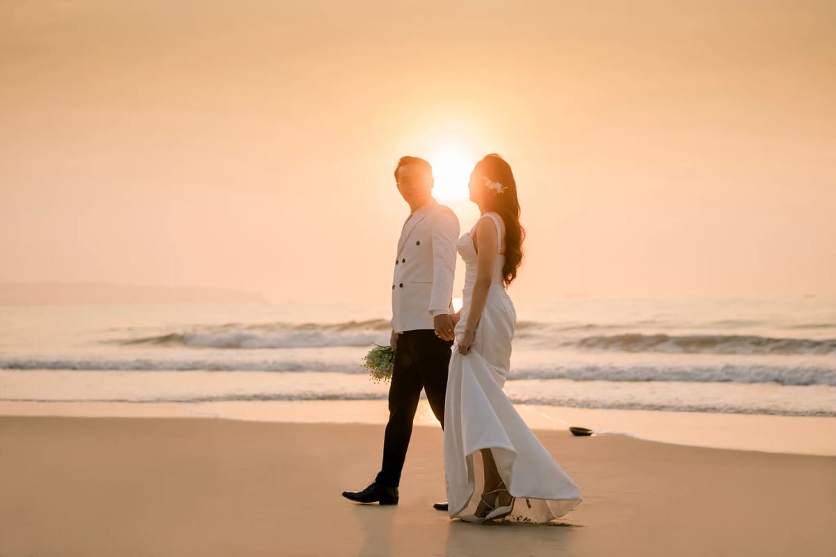 Cặp đôi bước đi trên bãi cát ướt ngược nắng hoàng hôn, cô dâu tay nâng tà váy dài