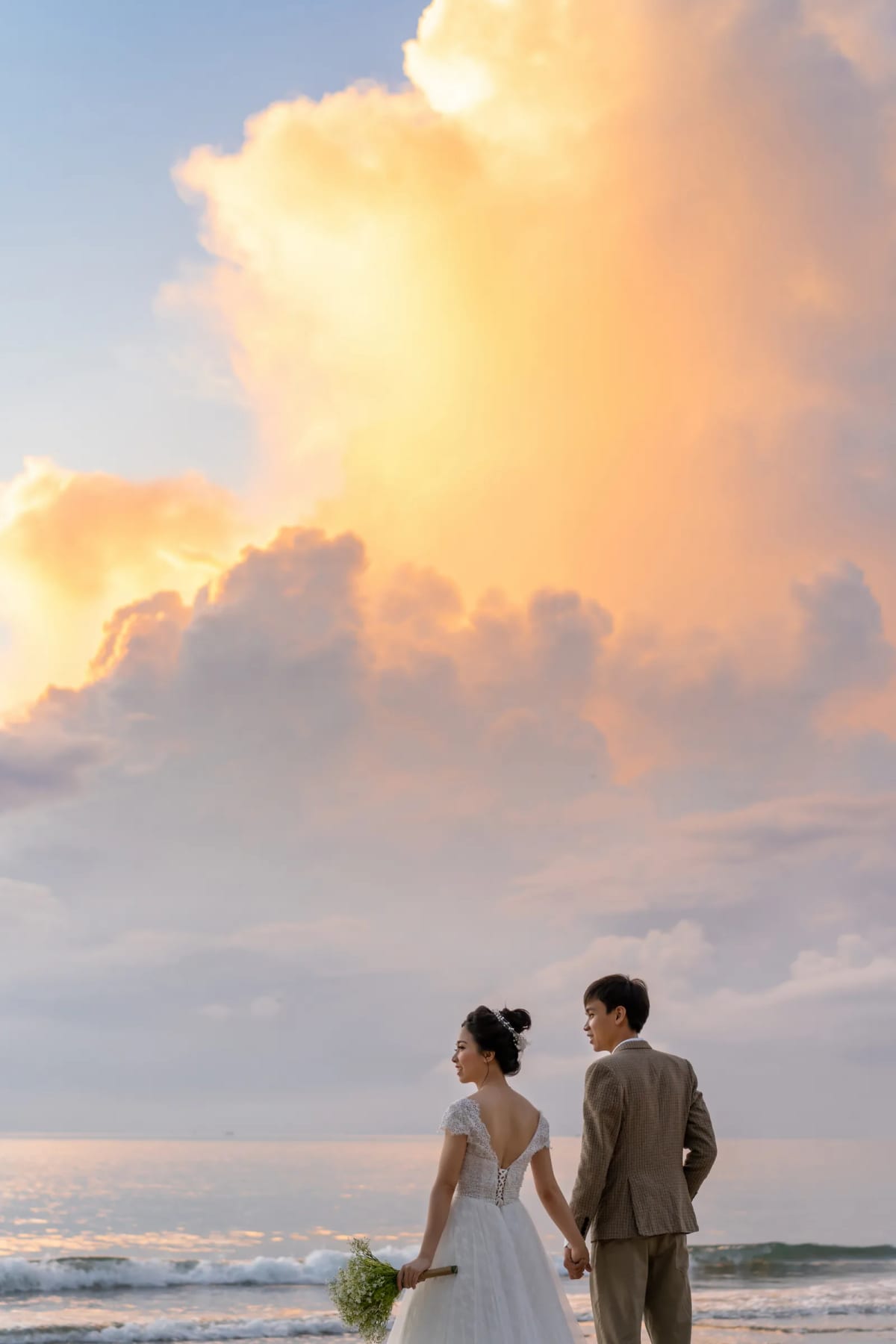 Cô dâu chú rể nắm tay đứng bên bờ sóng, phía trên là khối mây lớn nhuộm vàng cam chiếm gần hết khung hình