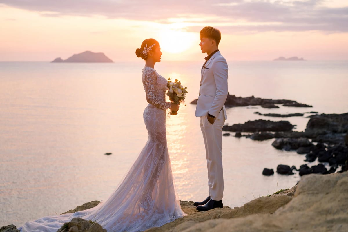 Cô dâu váy ren dài và chú rể vest trắng đứng đối diện trên mỏm đá, mặt trời lặn sau lưng in vệt vàng xuống biển