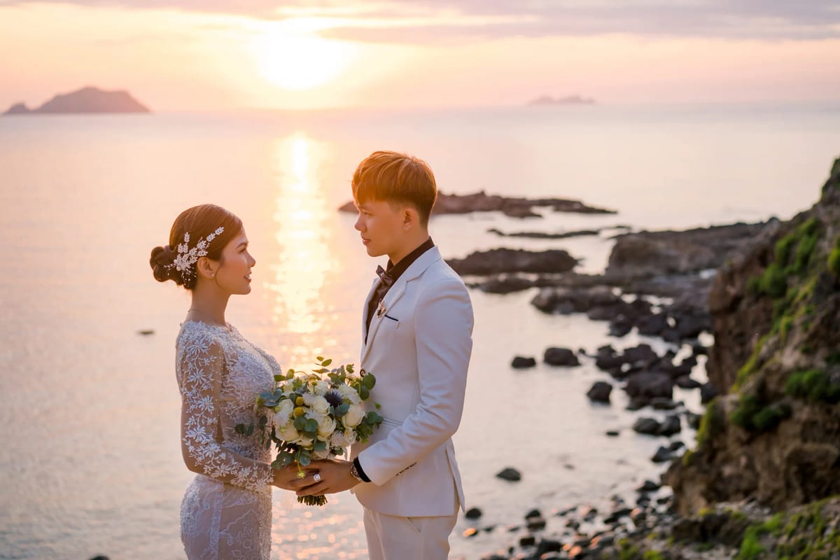 Cận cảnh cô dâu chú rể cùng cầm bó hoa cưới, phía sau là mặt biển ánh hồng lúc mặt trời xuống