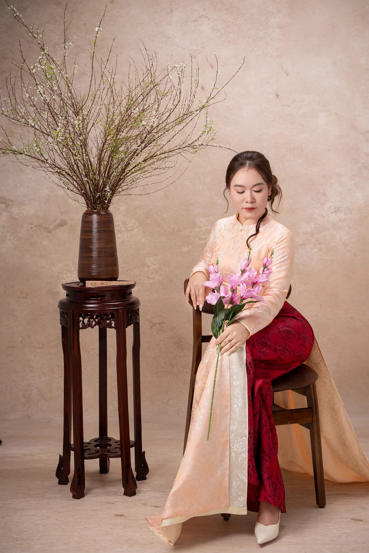 Cô gái áo dài cam ngồi trên ghế gỗ, tay ôm bó hoa lay ơn hồng, bên cạnh là bình hoa mai trắng trên đôn gỗ