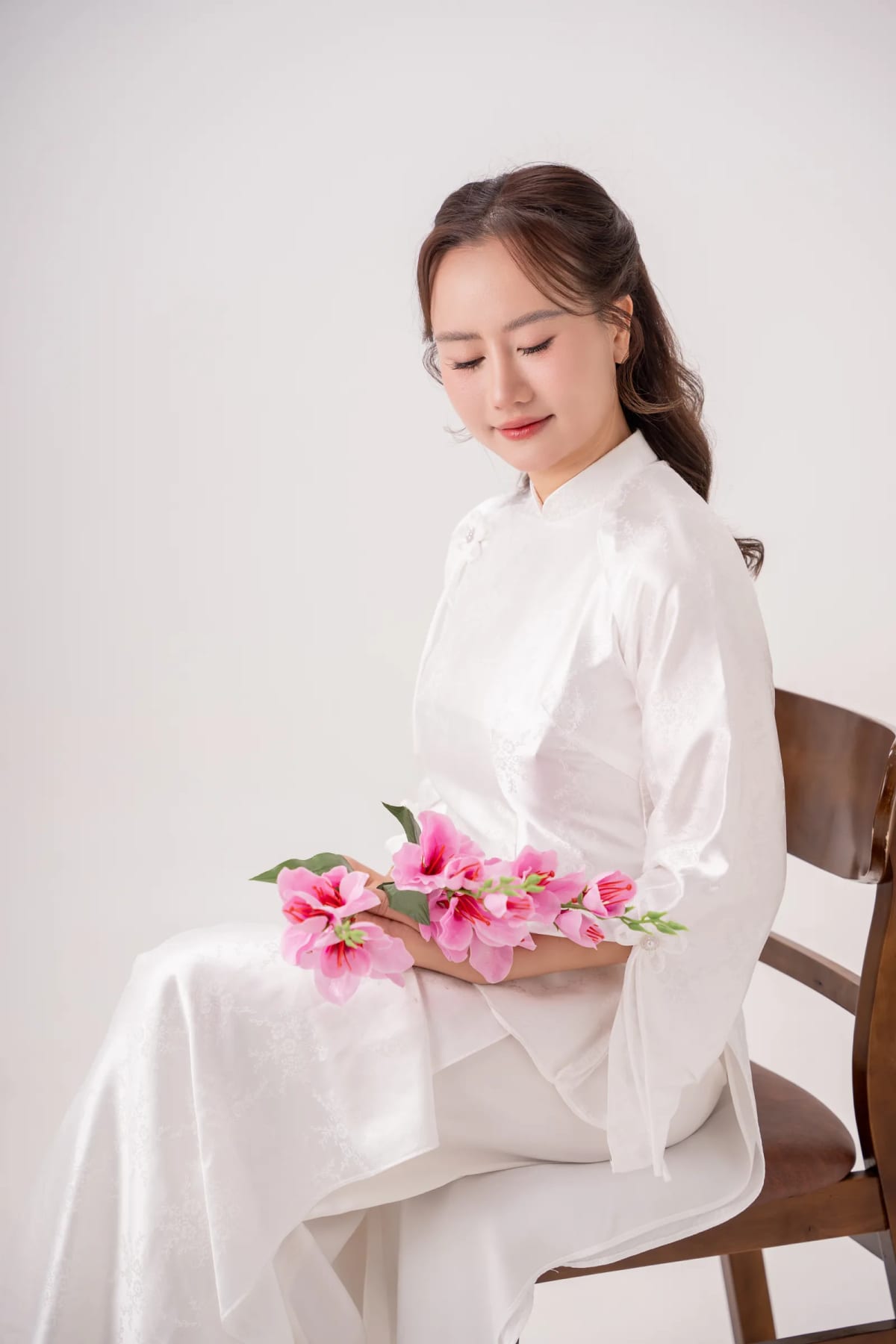 Cô gái áo dài trắng ngồi trên ghế gỗ, cúi nhìn bó hoa hồng đặt trên đùi