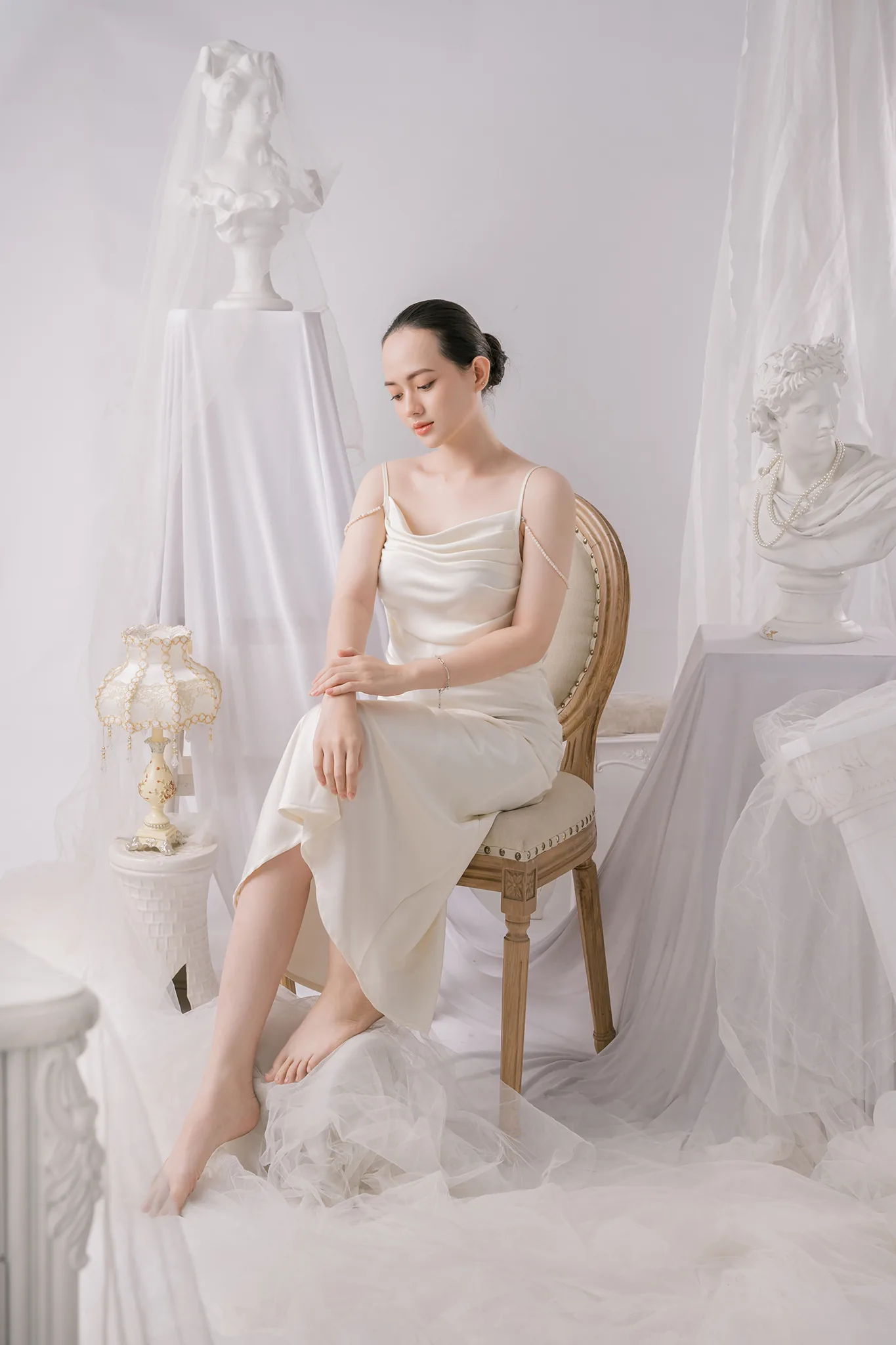 Cô gái mặc váy lụa trắng ngồi trên ghế, xung quanh là tượng thạch cao và vải voan trắng