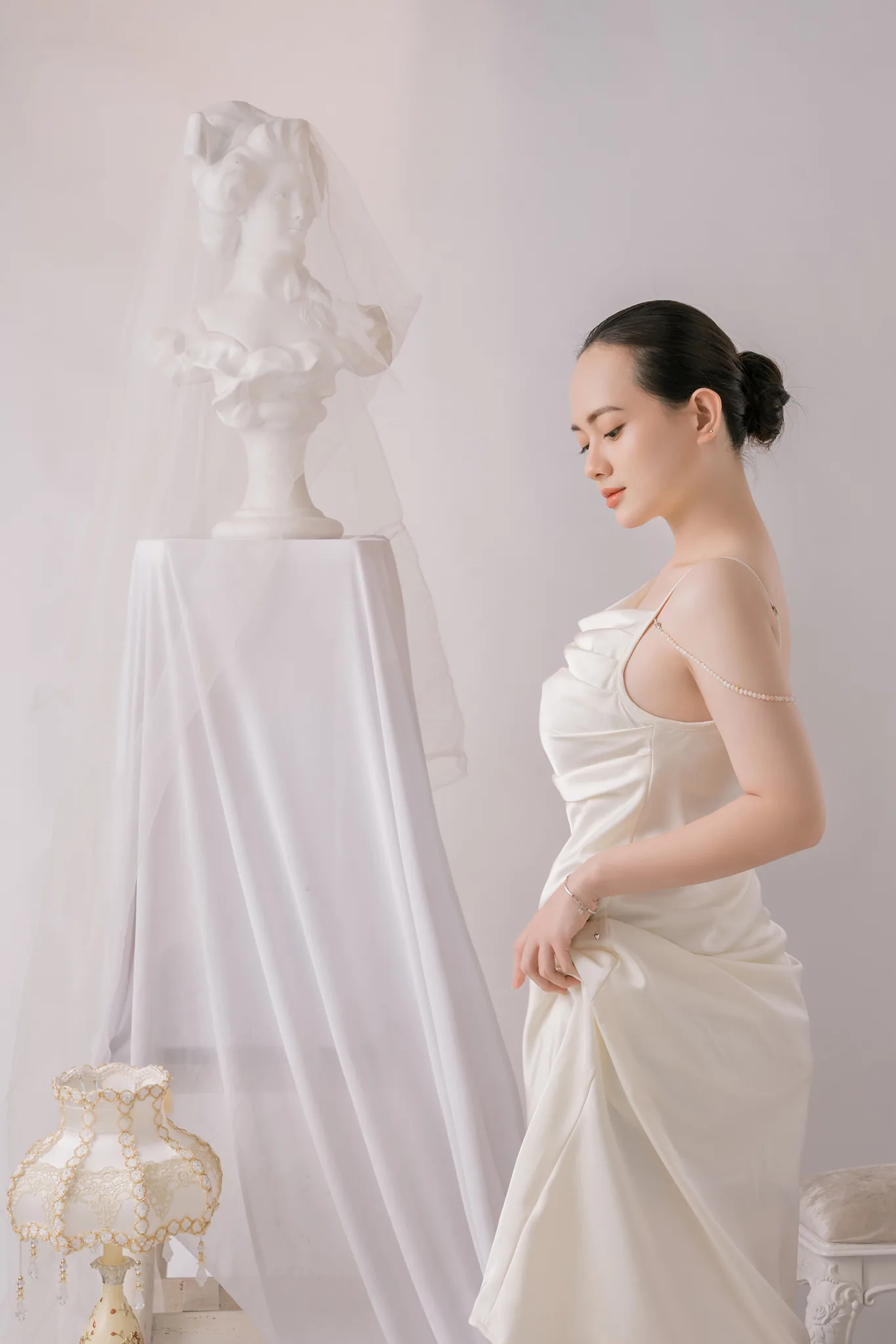Cô gái váy lụa trắng đứng nghiêng, tay nâng nhẹ tà váy, bên tượng bán thân phủ voan