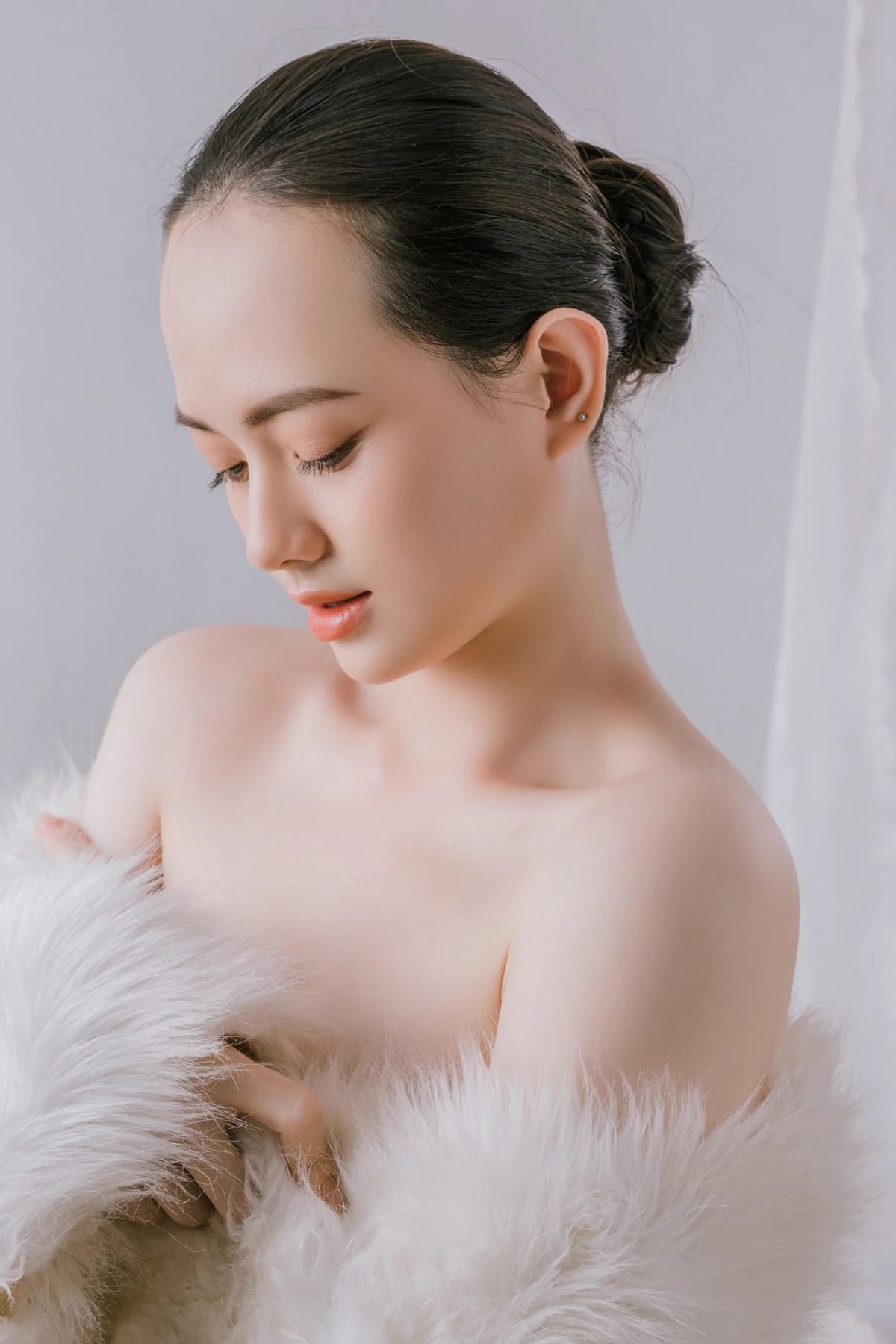 Chân dung cận cảnh cô gái búi tóc gọn, khoác lông vũ trắng, cúi mặt trong ánh sáng dịu