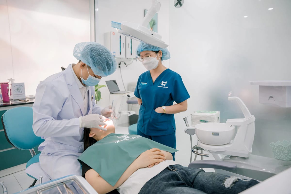 Bác sĩ nha khoa đang thăm khám cho bệnh nhân trên ghế nha, trợ thủ mặc áo xanh đứng quan sát bên cạnh