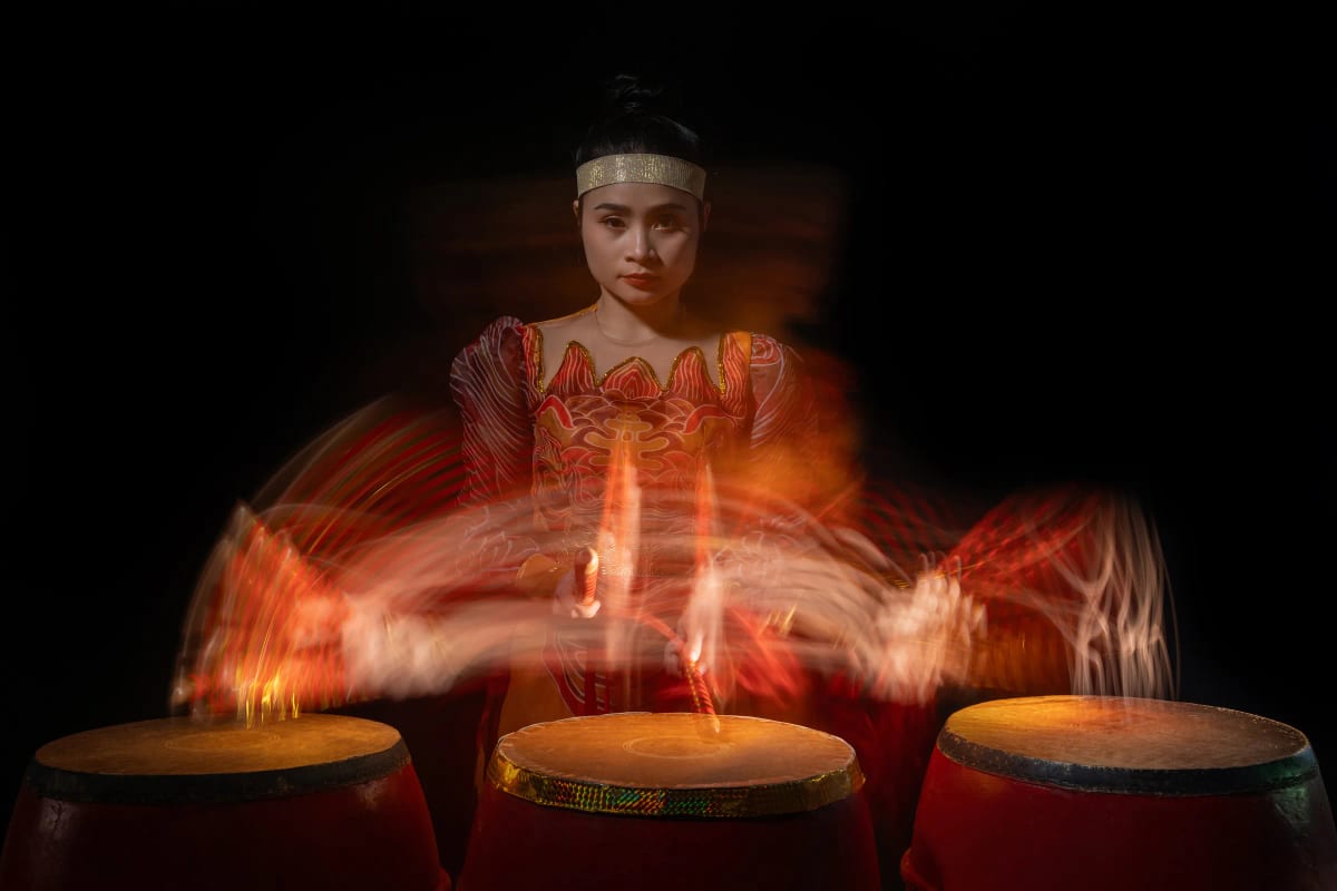 Người phụ nữ mặc áo đỏ đánh trống trận, phơi sáng dài biến hai cánh tay thành những vòng cung ánh sáng