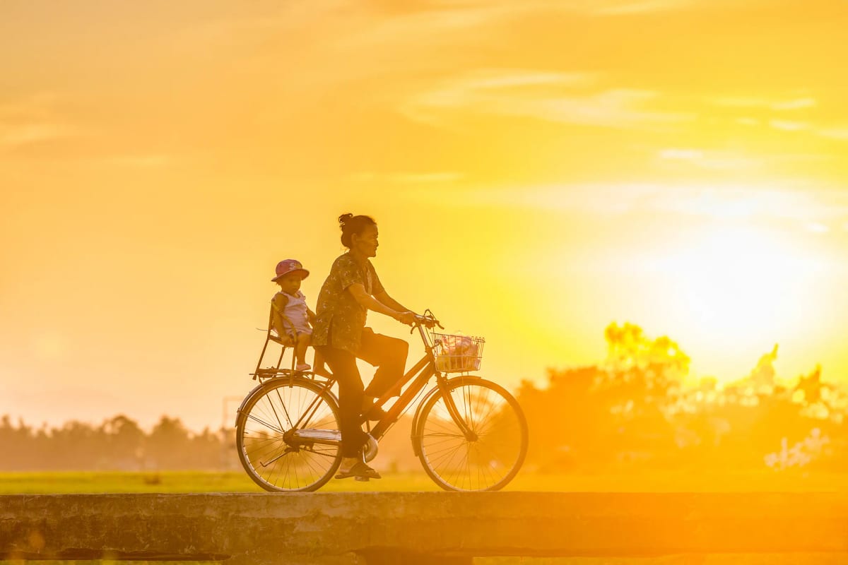 Người bà chở cháu nhỏ trên xe đạp băng qua bờ ruộng, cả hai in bóng trên nền trời vàng rực lúc hoàng hôn