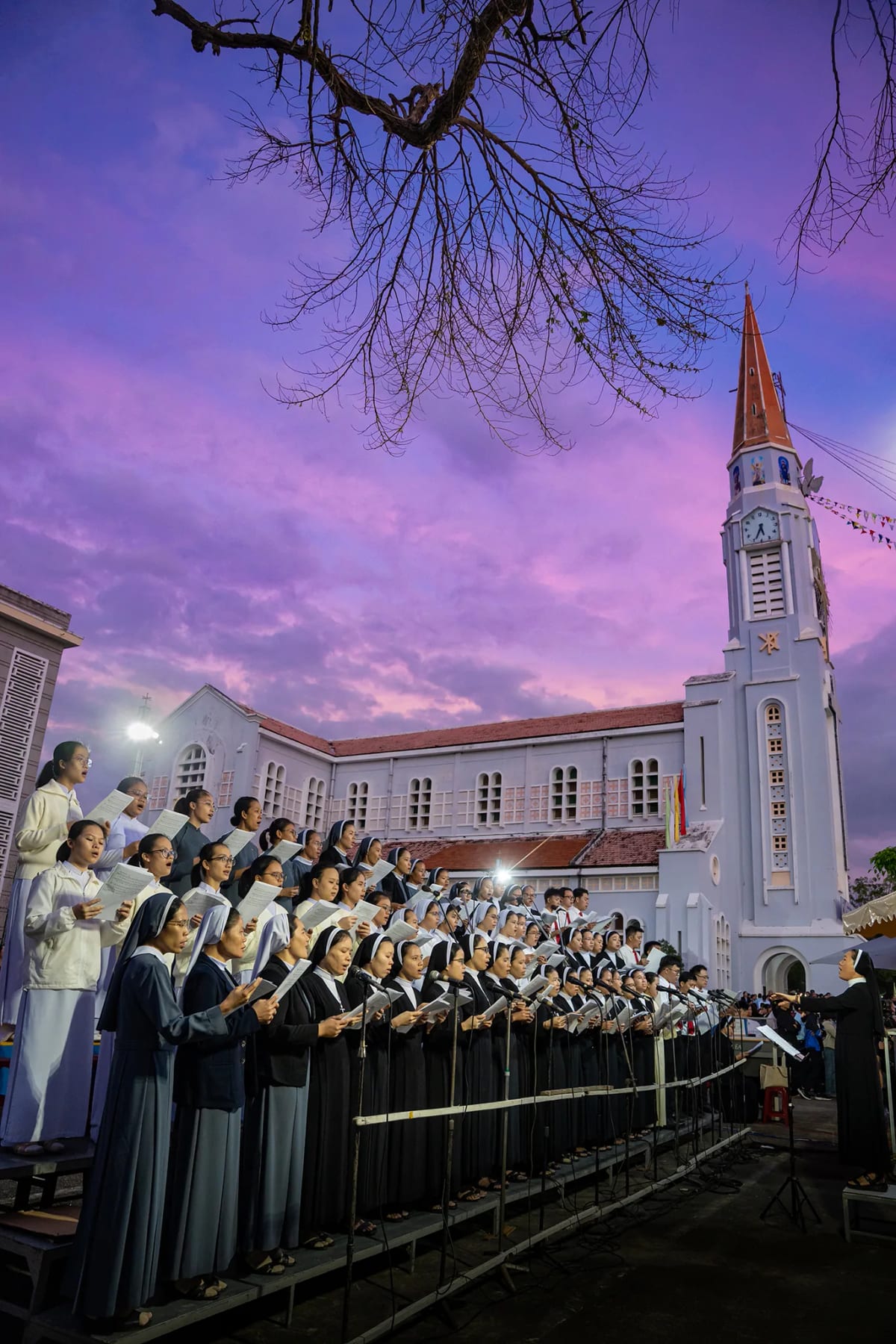 Ca đoàn hàng trăm nữ tu và giáo dân đứng hát trên khán đài trước nhà thờ trắng, trời hoàng hôn tím hồng, một cành cây khô vươn ngang khung hình