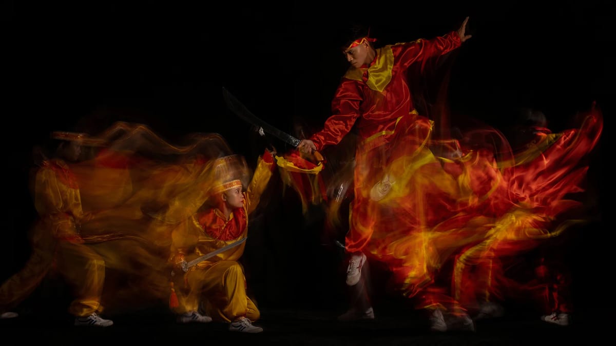Hai võ sinh giao đấu, người áo đỏ bật lên vung đao chém xuống, người áo vàng tấn thấp đỡ đòn, quanh họ là những dải lụa đỏ vàng