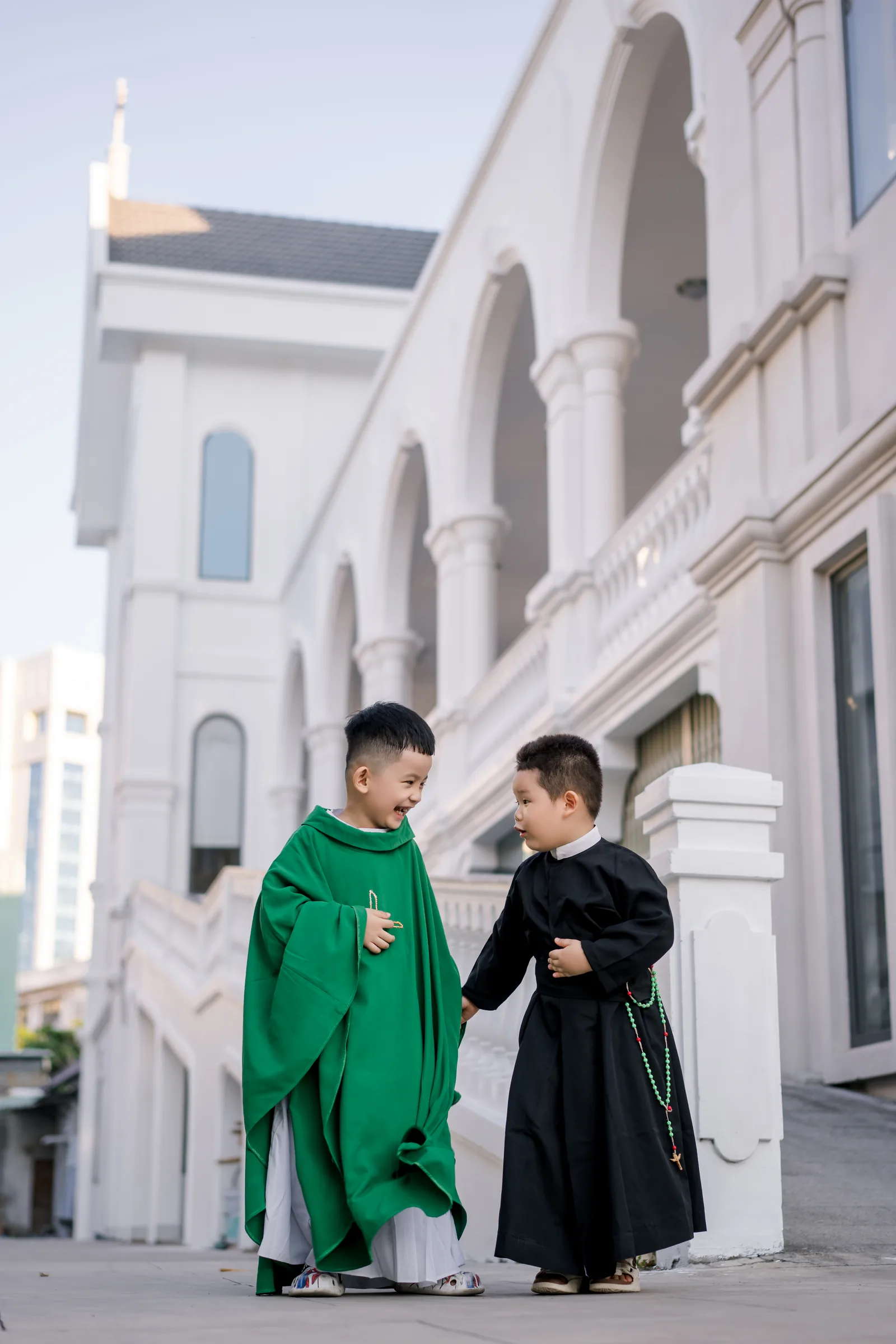 Hai cậu bé đứng trước nhà thờ trắng quay sang cười với nhau, một em mặc áo lễ xanh lá, một em mặc áo chùng thâm đeo chuỗi hạt xanh