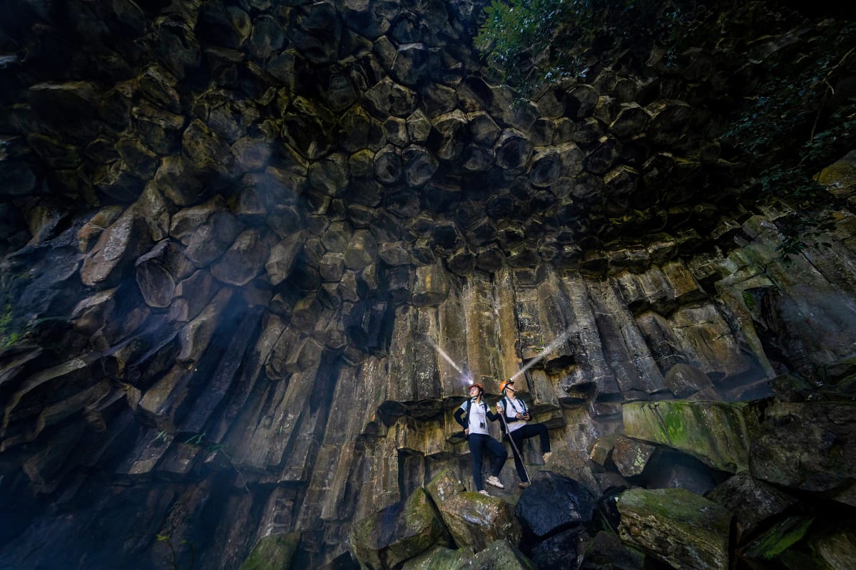 Hai người đội mũ bảo hộ đeo đèn pin đứng nhỏ bé dưới vòm đá bazan hình lục giác khổng lồ ở thác Hang Dơi
