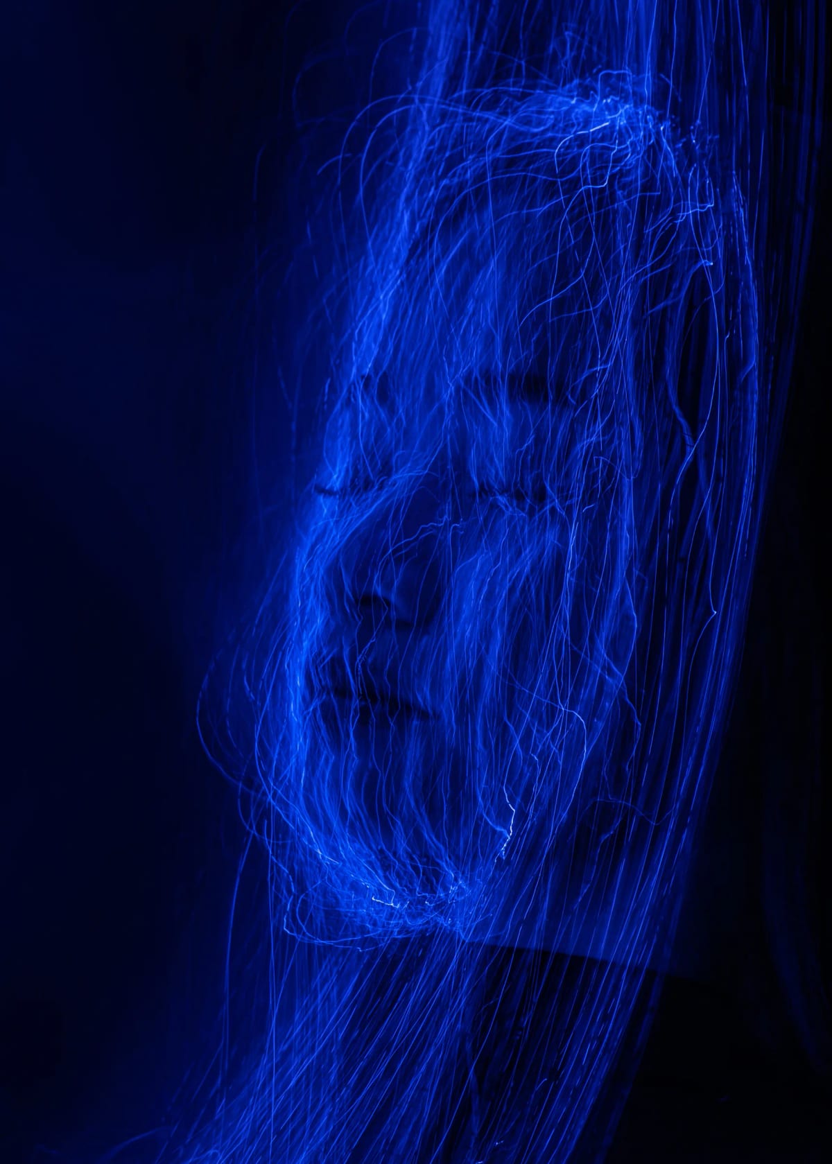 Chân dung phơi sáng dài, gương mặt chìm dưới hàng nghìn sợi sáng xanh dương đan chéo trên nền đen