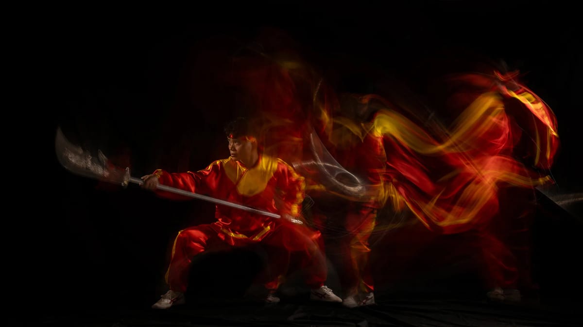 Võ sinh áo đỏ viền vàng tấn thấp chĩa đại đao ra trước, chuyển động phía sau kéo thành những vệt lụa đỏ cuộn như mây lửa