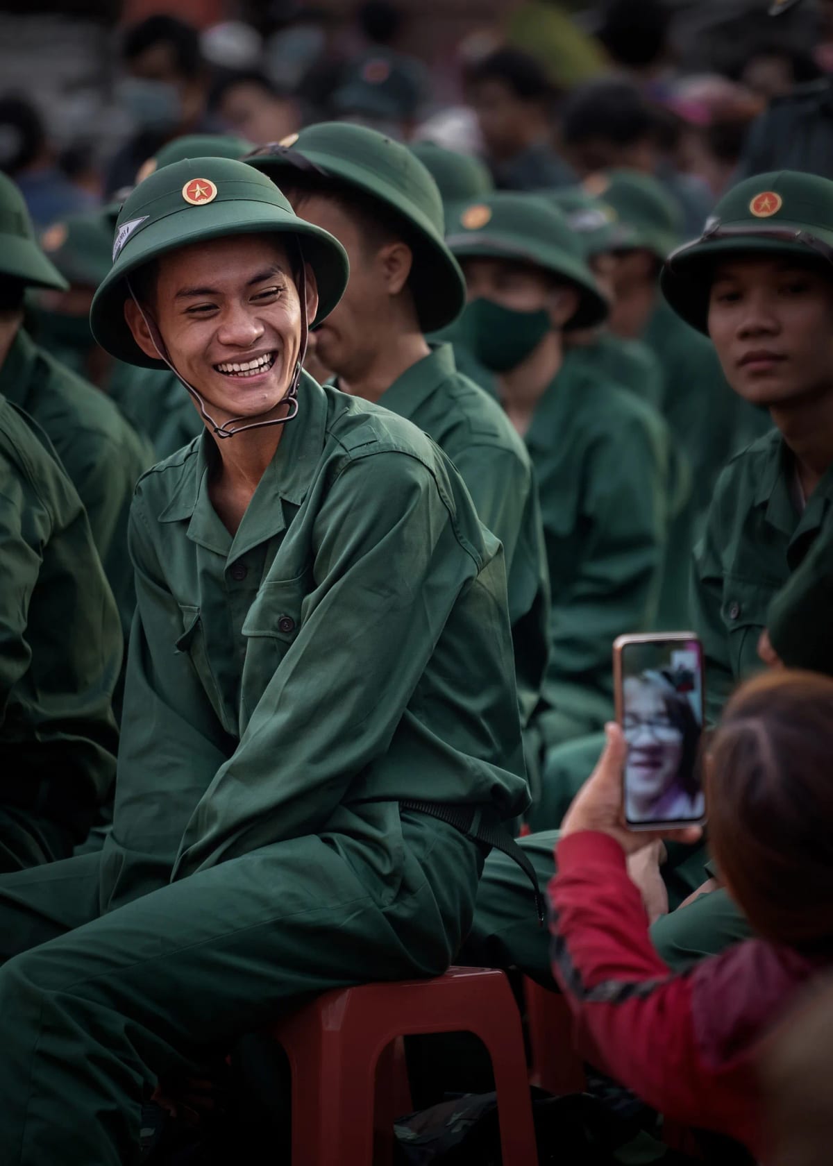 Tân binh mặc quân phục xanh đội mũ cối gắn sao vàng quay lại cười rạng rỡ, phía trước một người thân giơ điện thoại đang gọi video