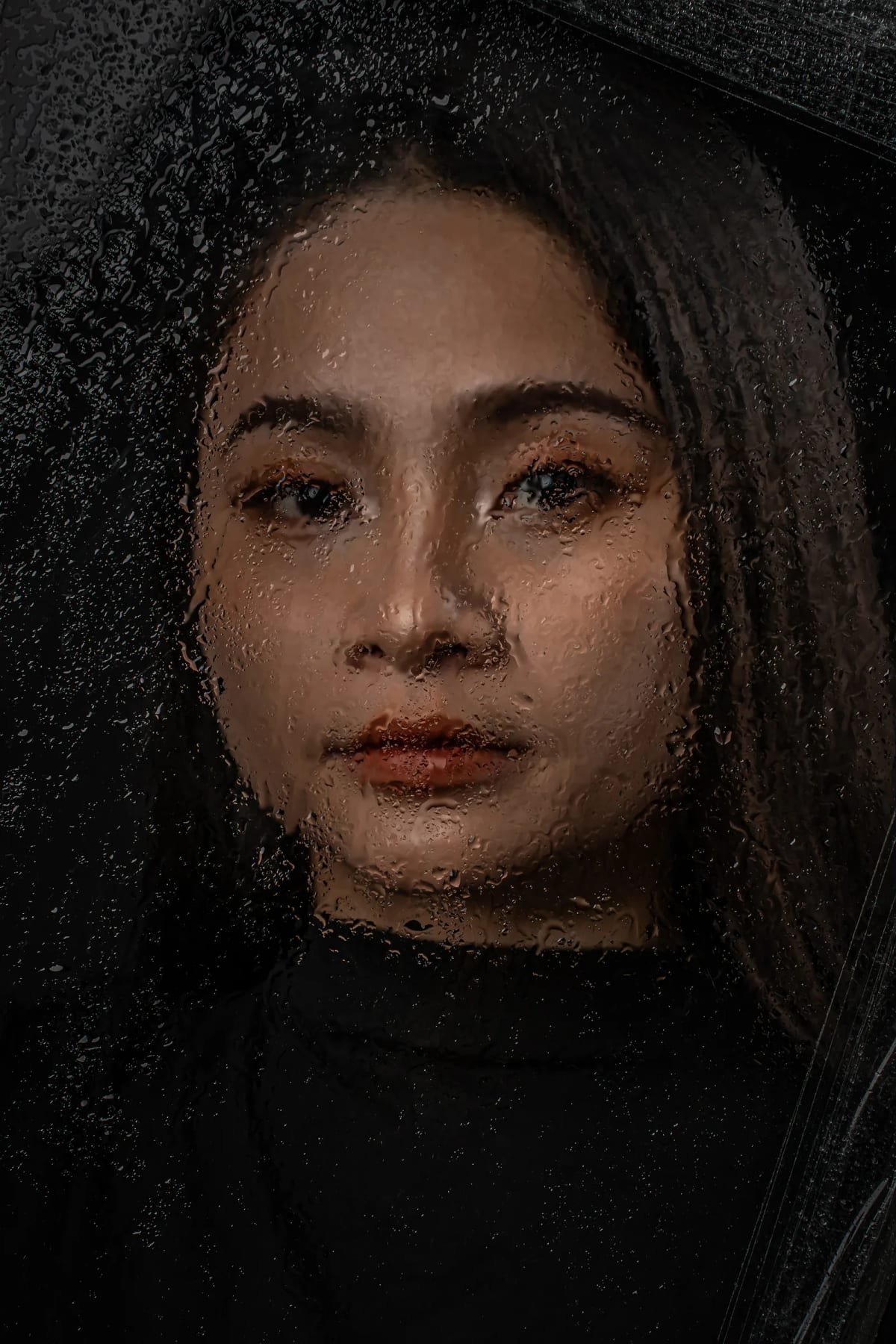 Gương mặt cô gái nhìn qua tấm kính phủ đầy hạt nước, xung quanh tối đen, chỉ đôi mắt và làn môi còn rõ nét