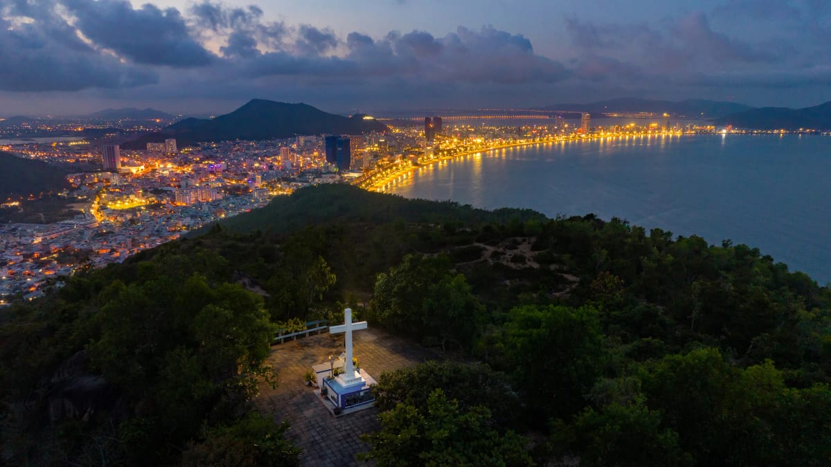 Ảnh flycam cây thánh giá trắng trên đỉnh đồi cây xanh, phía xa là vịnh biển và dải đèn thành phố lúc trời chạng vạng