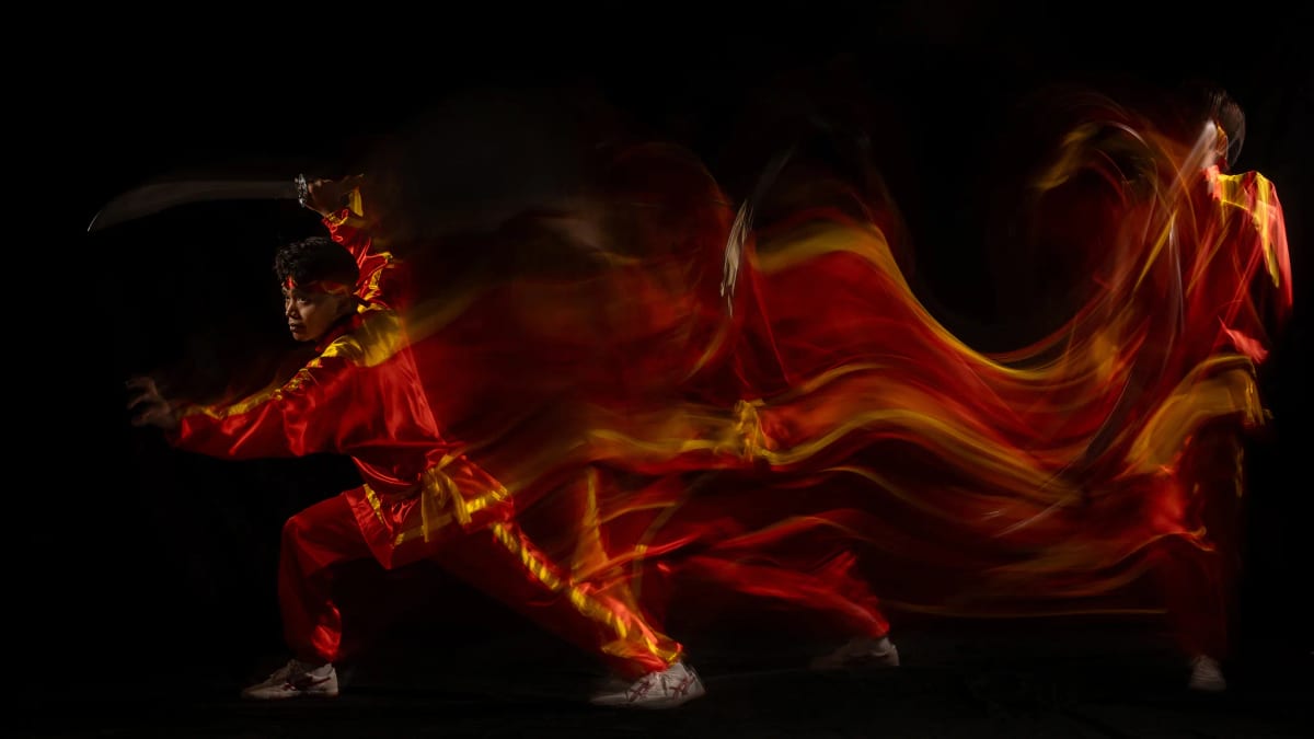 Võ sinh áo đỏ vung siêu đao ngang đầu ở thế tấn thấp, những vệt lụa đỏ và vàng loang phía sau thành hình một con thú đang lao tới