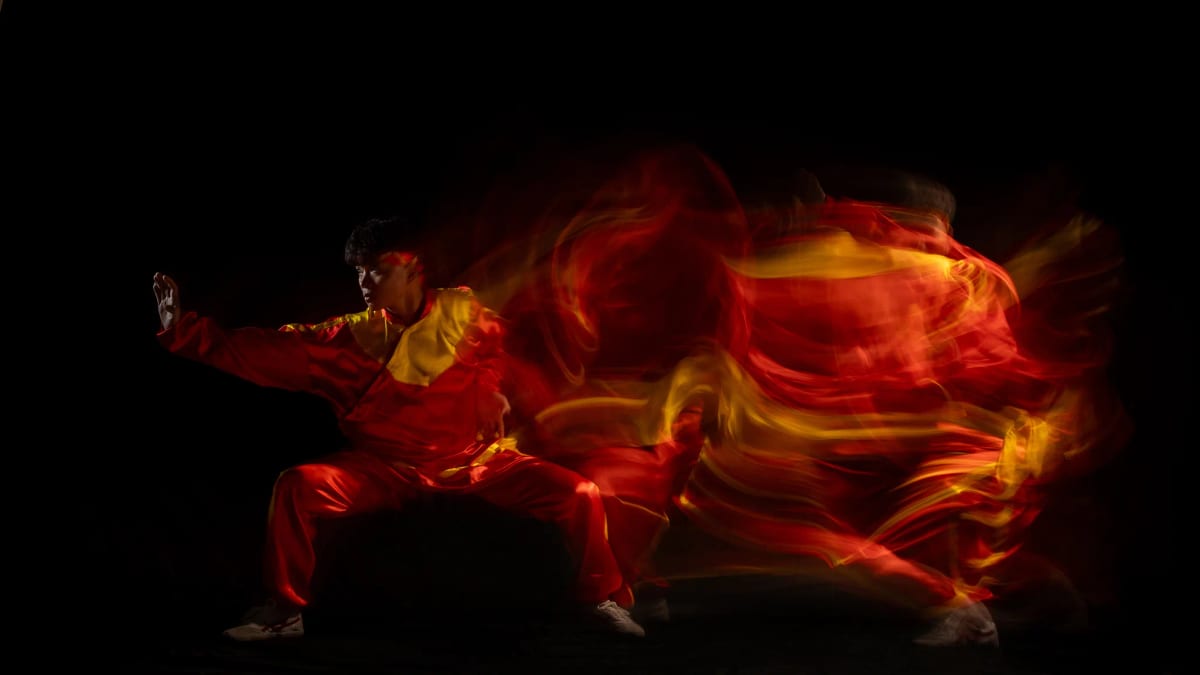 Võ sinh áo đỏ vàng tấn ngang xoè tay trái ra trước, nửa thân sau tan thành những dải lụa đỏ và vàng cuộn xoáy