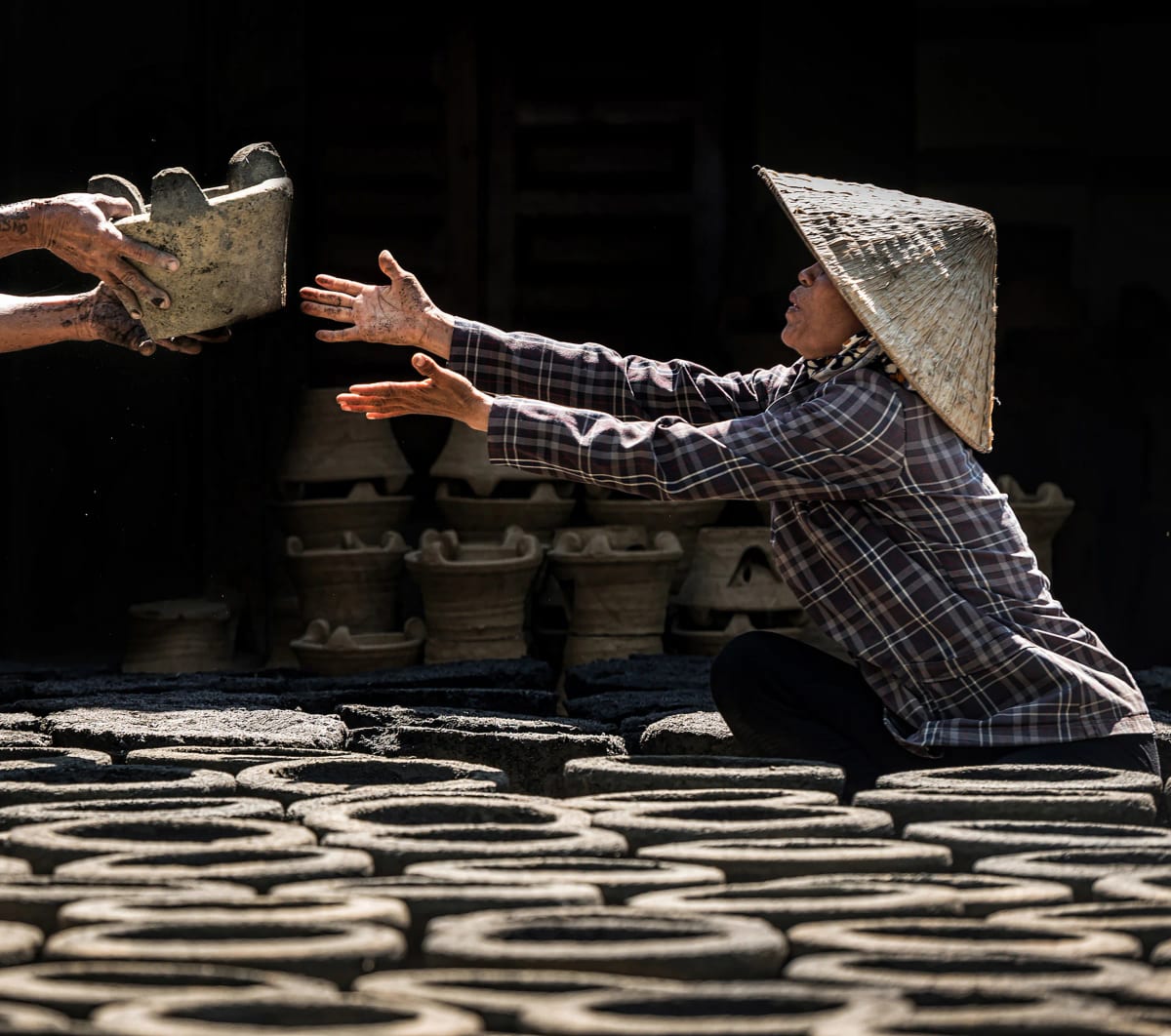 Người phụ nữ đội nón lá quỳ giữa sân phơi, vươn hai tay đón chiếc lò đất từ đôi tay người đối diện, phía sau là hàng lò gốm mộc xếp chồng