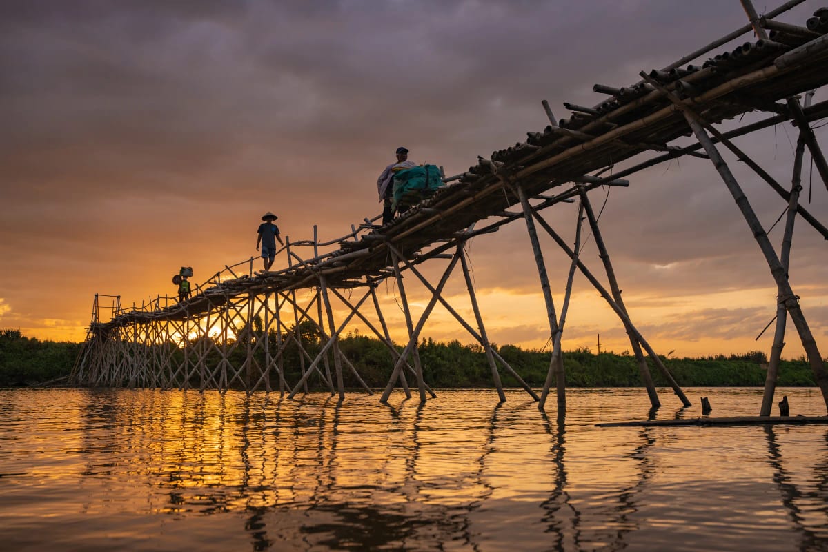 Ba người gánh hàng đi trên chiếc cầu tre cao lênh khênh vắt qua sông, trời chiều tím và cam in bóng xuống mặt nước