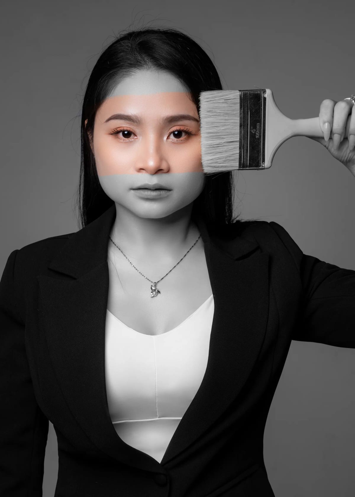Cô gái mặc vest đen giơ chiếc cọ sơn bản lớn ngang mặt, nửa khuôn mặt và thân người đã bị quét thành màu xám trắng, nửa còn lại vẫn nguyên màu da