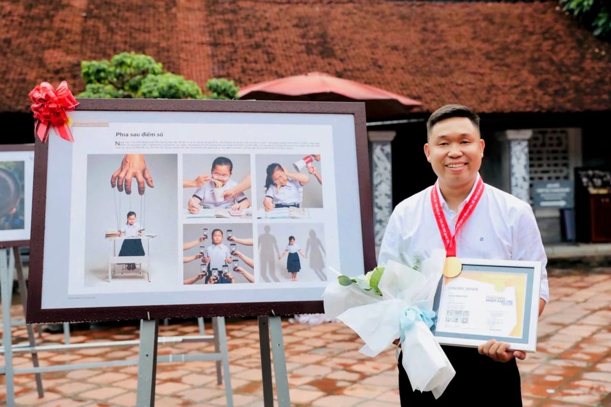 Phan Minh Thọ đeo huy chương, cầm hoa và bằng chứng nhận, đứng cạnh bảng trưng bày ảnh bộ Phía sau điểm số gồm năm khung tại Văn Miếu