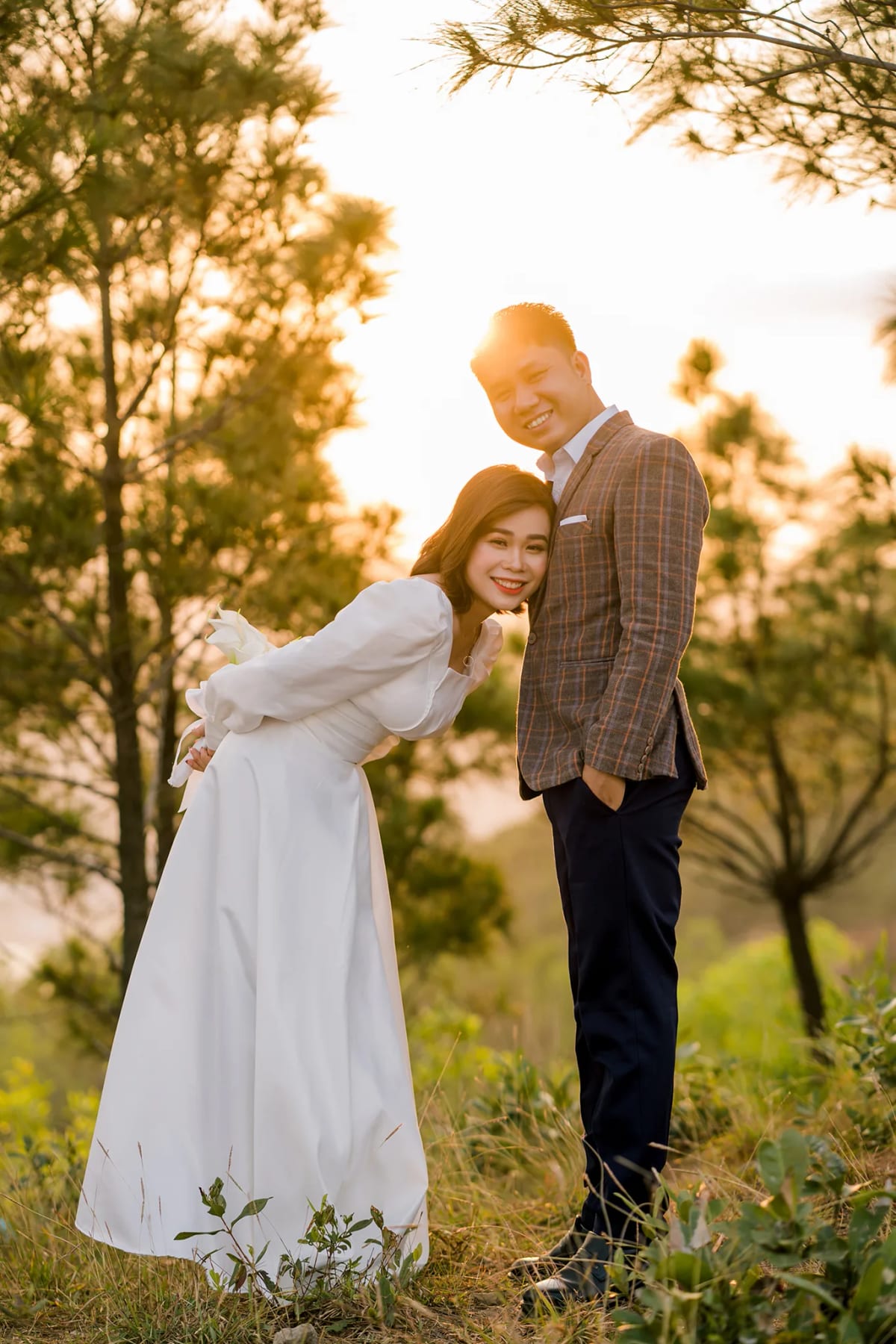 Cô dâu váy trắng ngả người tựa vào chú rể trong rừng thông ngược nắng chiều