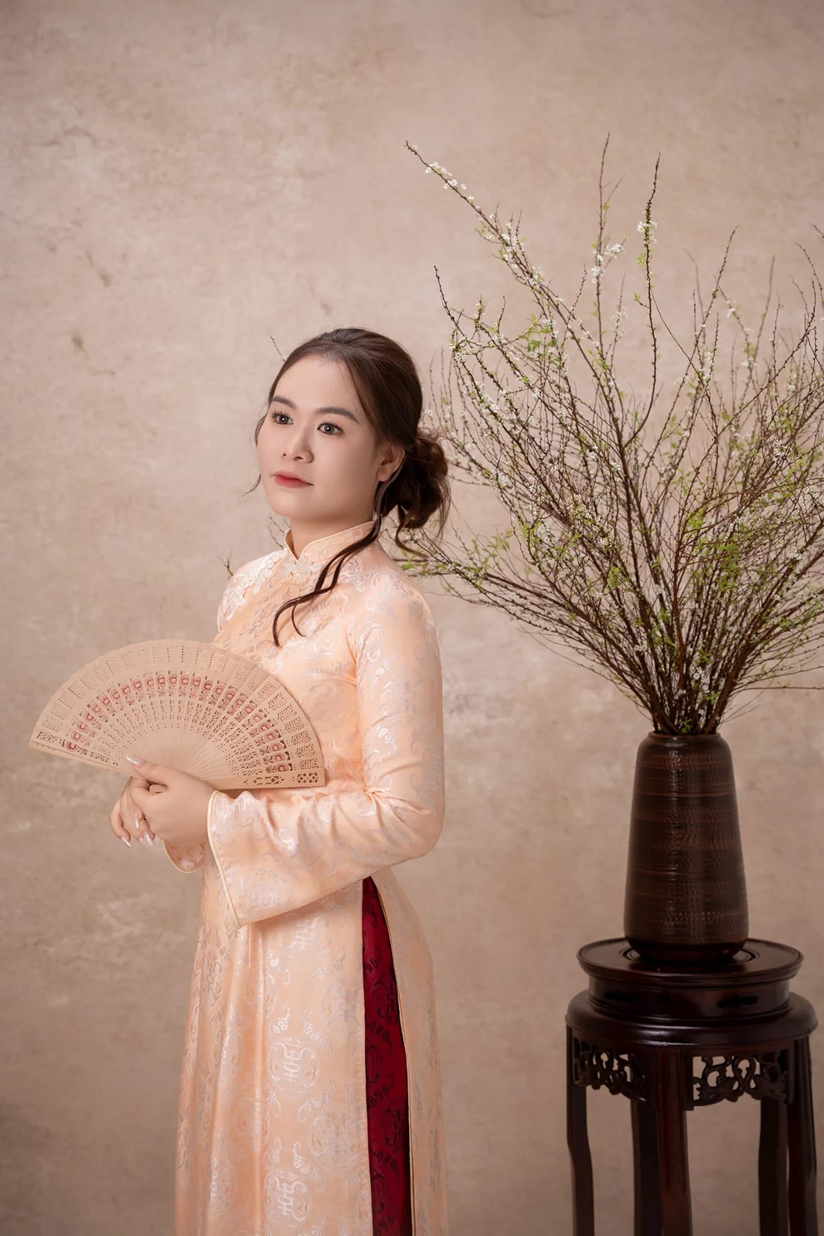Cô gái mặc áo dài gấm màu cam nhạt cầm quạt giấy, đứng bên bình hoa mai trắng