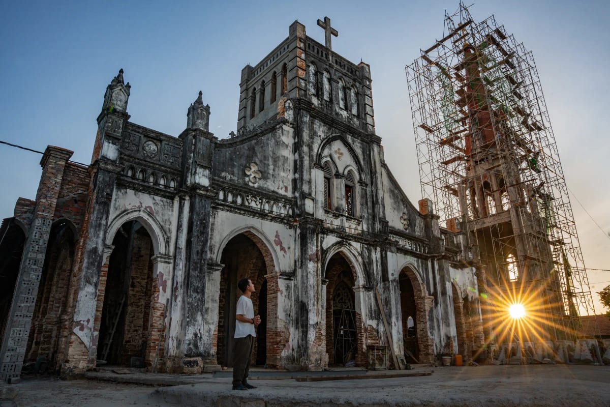 Người đàn ông áo trắng chắp tay ngước nhìn mặt tiền nhà thờ cổ tróc vữa, mặt trời lặn xé thành tia sáng sau giàn giáo tháp chuông